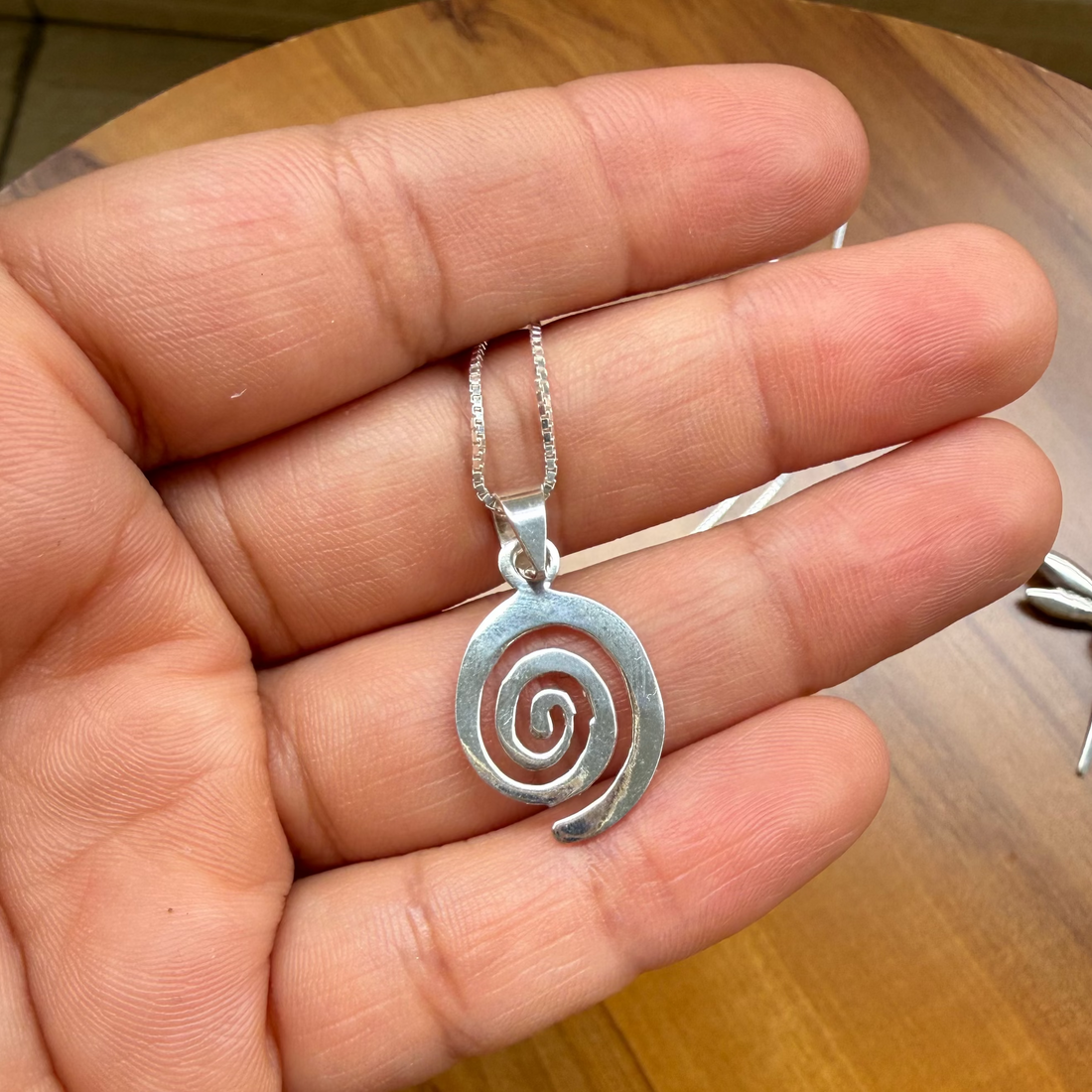 A hand holding a silver spiral pendant necklace on a wooden table background.