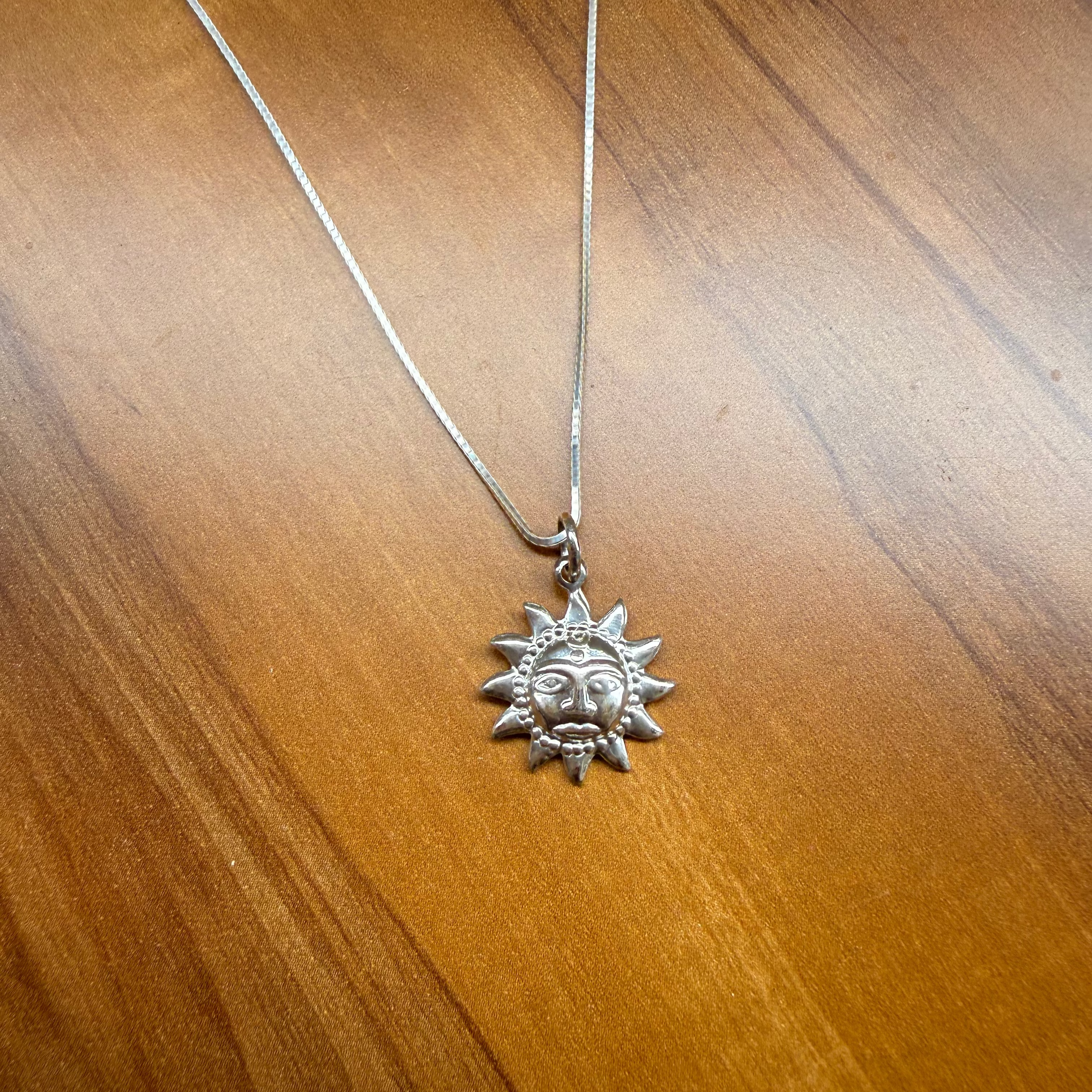 A silver sun pendant necklace with a detailed face and rays, hanging from a matching silver chain on a wooden surface.