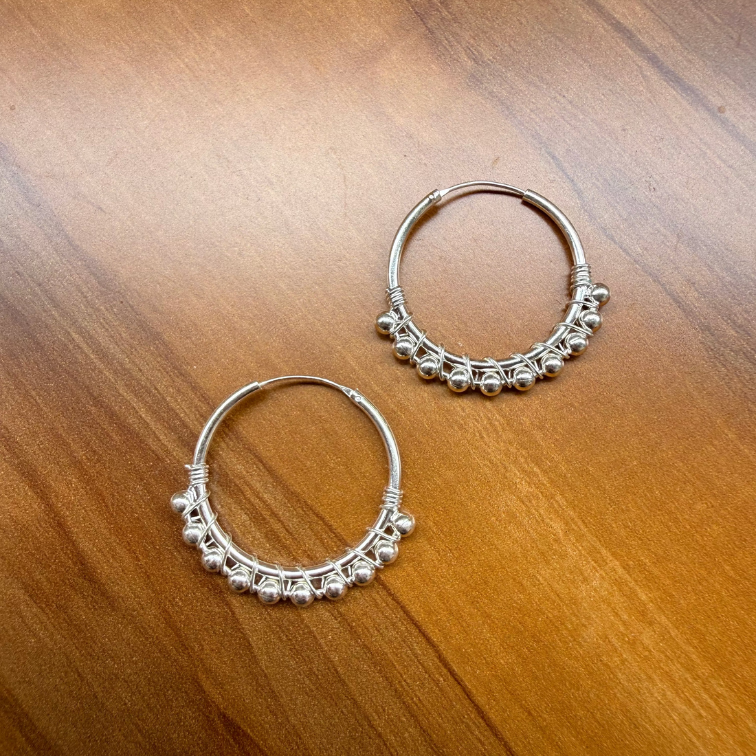 A pair of silver-toned hoop earrings with delicate beadwork along the lower curve, displayed on a warm brown wooden surface.
