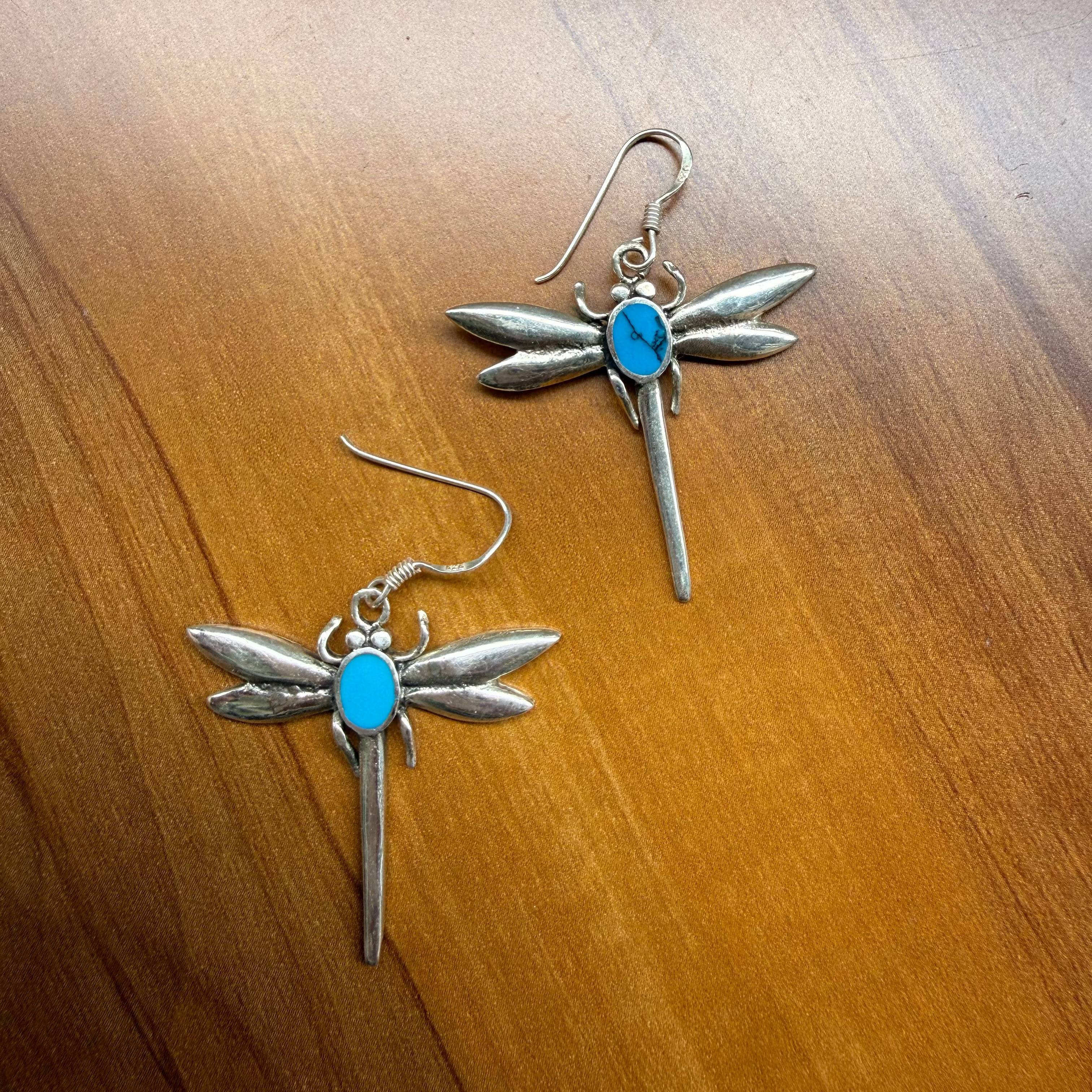 A pair of silver dragonfly earrings with bright blue turquoise centers, displayed on a warm brown wooden surface.