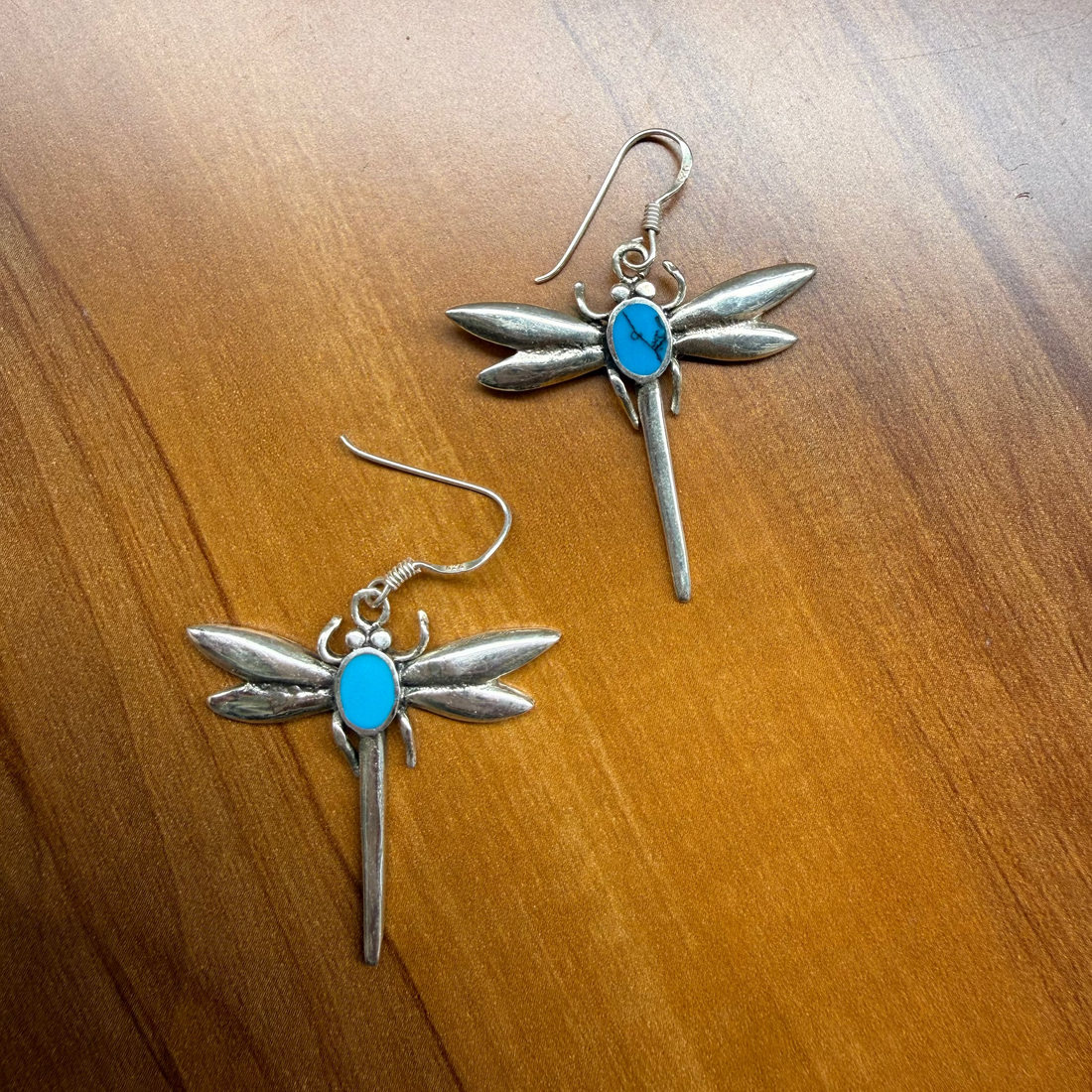 A pair of silver dragonfly earrings with bright blue turquoise centers, displayed on a warm brown wooden surface.