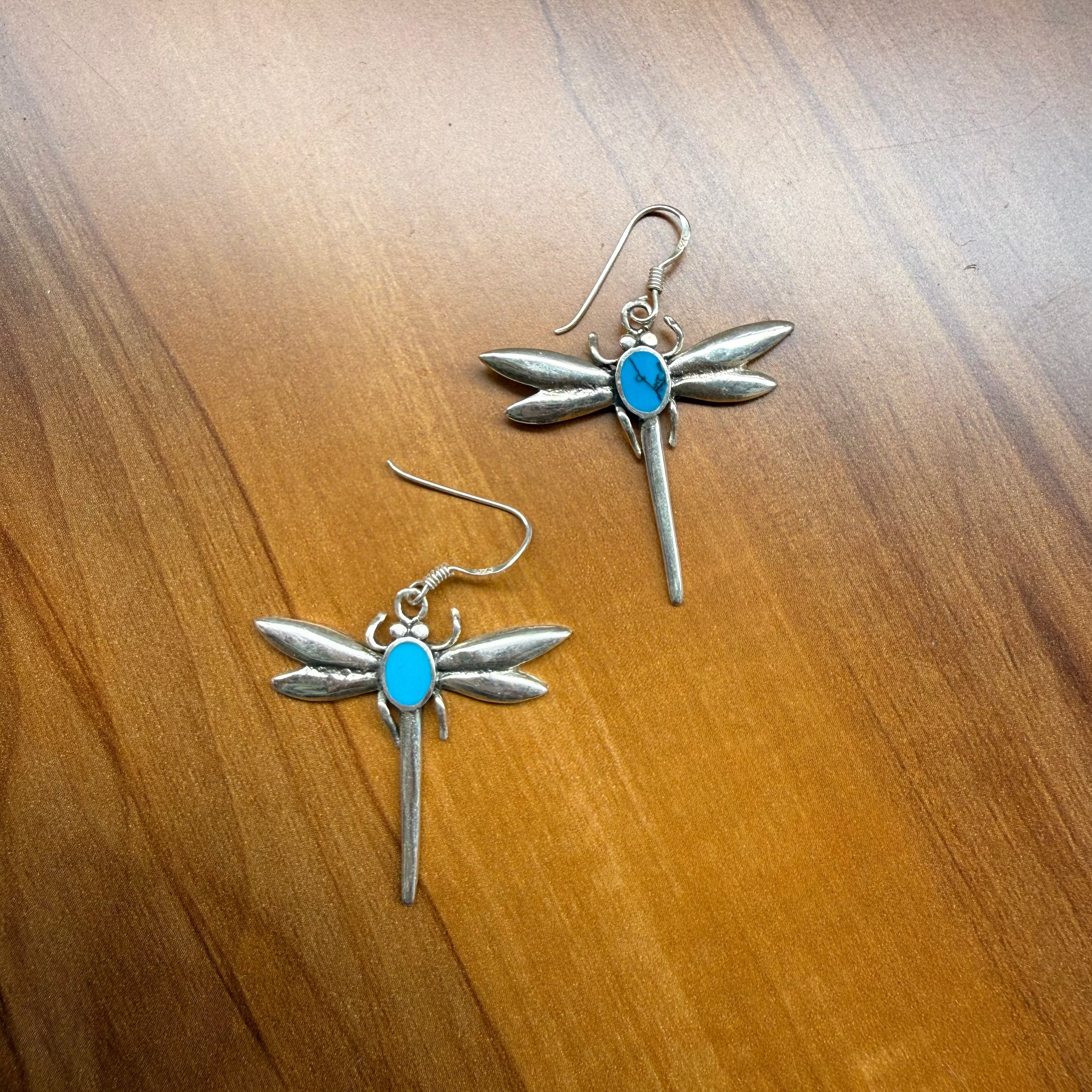 A pair of dragonfly-shaped earrings with silver-toned metal wings and a small oval turquoise blue stone in the center, displayed on a warm brown wooden surface.