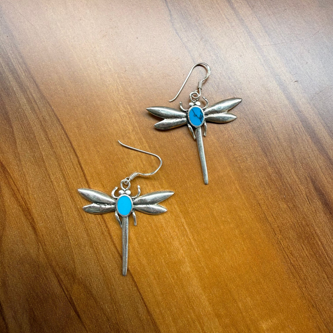 A pair of dragonfly-shaped earrings with silver-toned metal wings and a small oval turquoise blue stone in the center, displayed on a warm brown wooden surface.