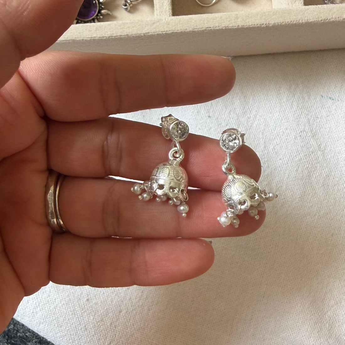 A hand holding a pair of silver-toned dangling earrings with small clear stones and dangling floral or leaf-like charms, with a jewelry tray in the background.