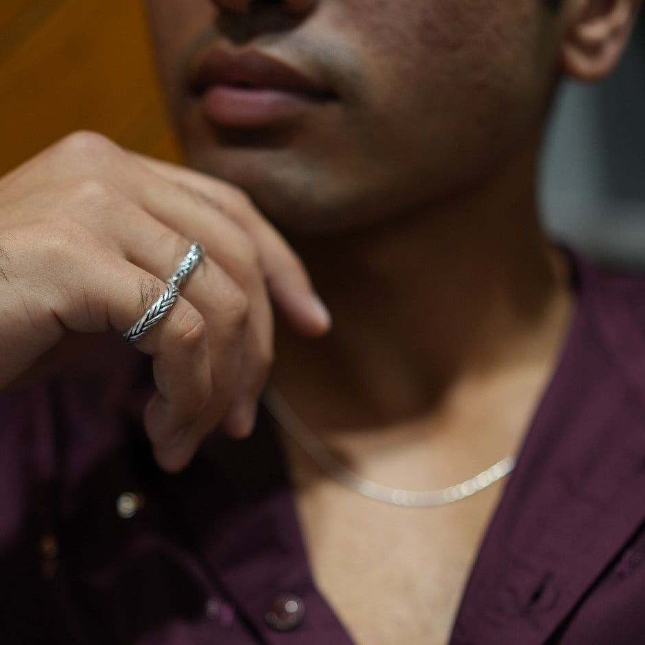 A close-up lifestyle shot of a man wearing a textured silver band ring on his finger, paired with a dark purple embroidered shirt and a silver chain necklace.