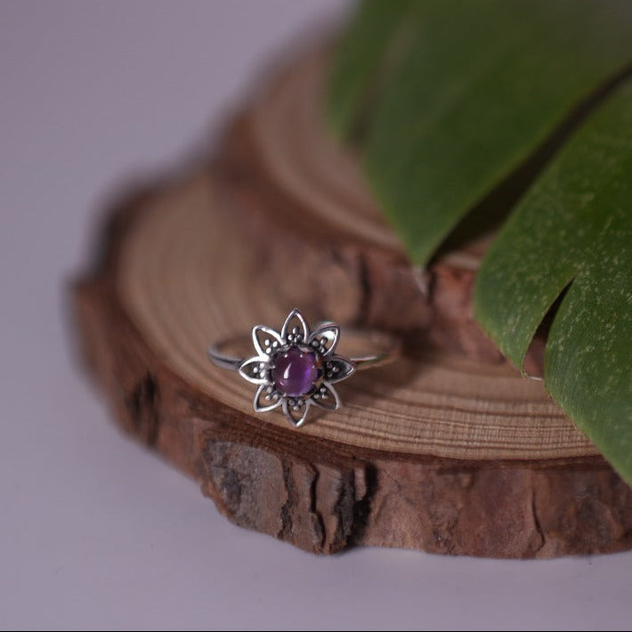 A silver ring with a flower-shaped setting and a small purple gemstone, displayed on a rustic wood slice with a green leaf against a soft light gray background.