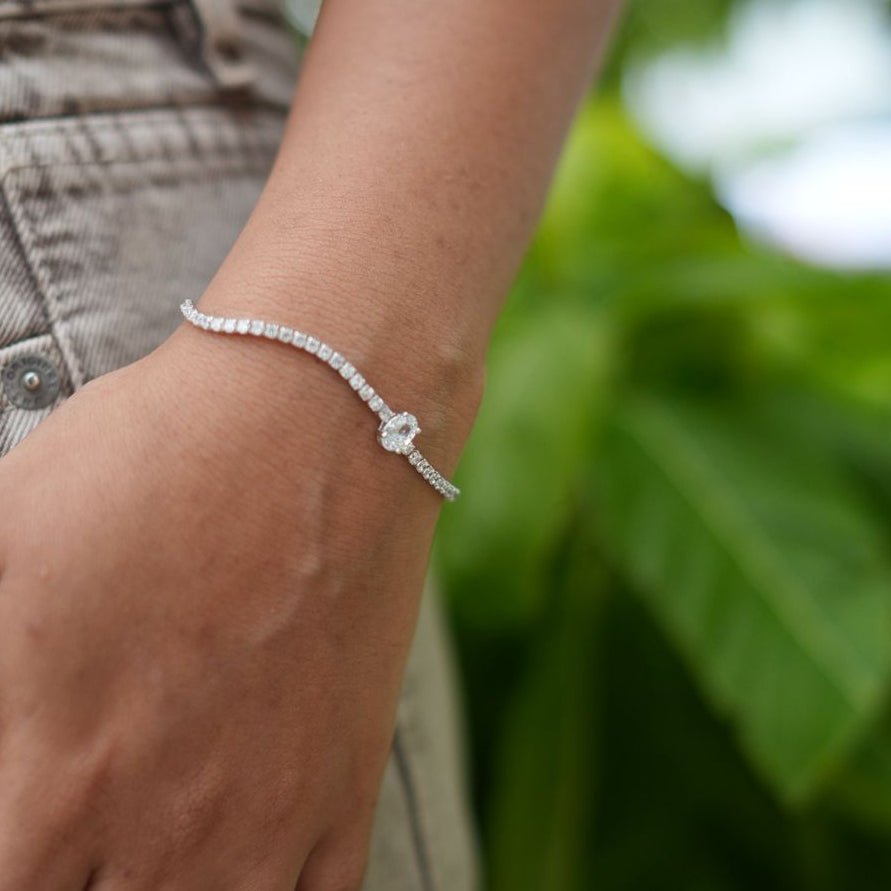 A close-up of a person's wrist wearing a delicate silver chain bracelet with a small heart-shaped charm, set against a soft-focus background of green leaves.