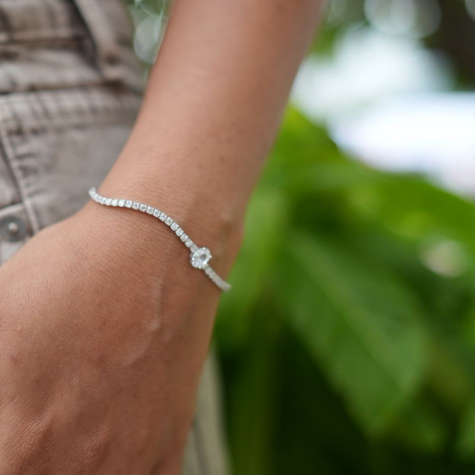 A delicate silver bracelet with a small central charm, worn on a person's wrist. The person is also wearing silver rings, and the background is a soft-focus outdoor scene with green foliage.