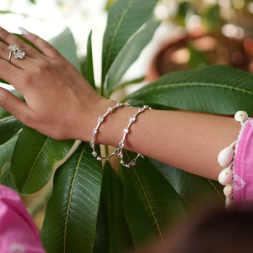 A close-up lifestyle shot of a person wearing silver jewelry, including a ring and bracelets, while gently touching the green leaves of a plant. The person is wearing a bright pink embroidered top.
