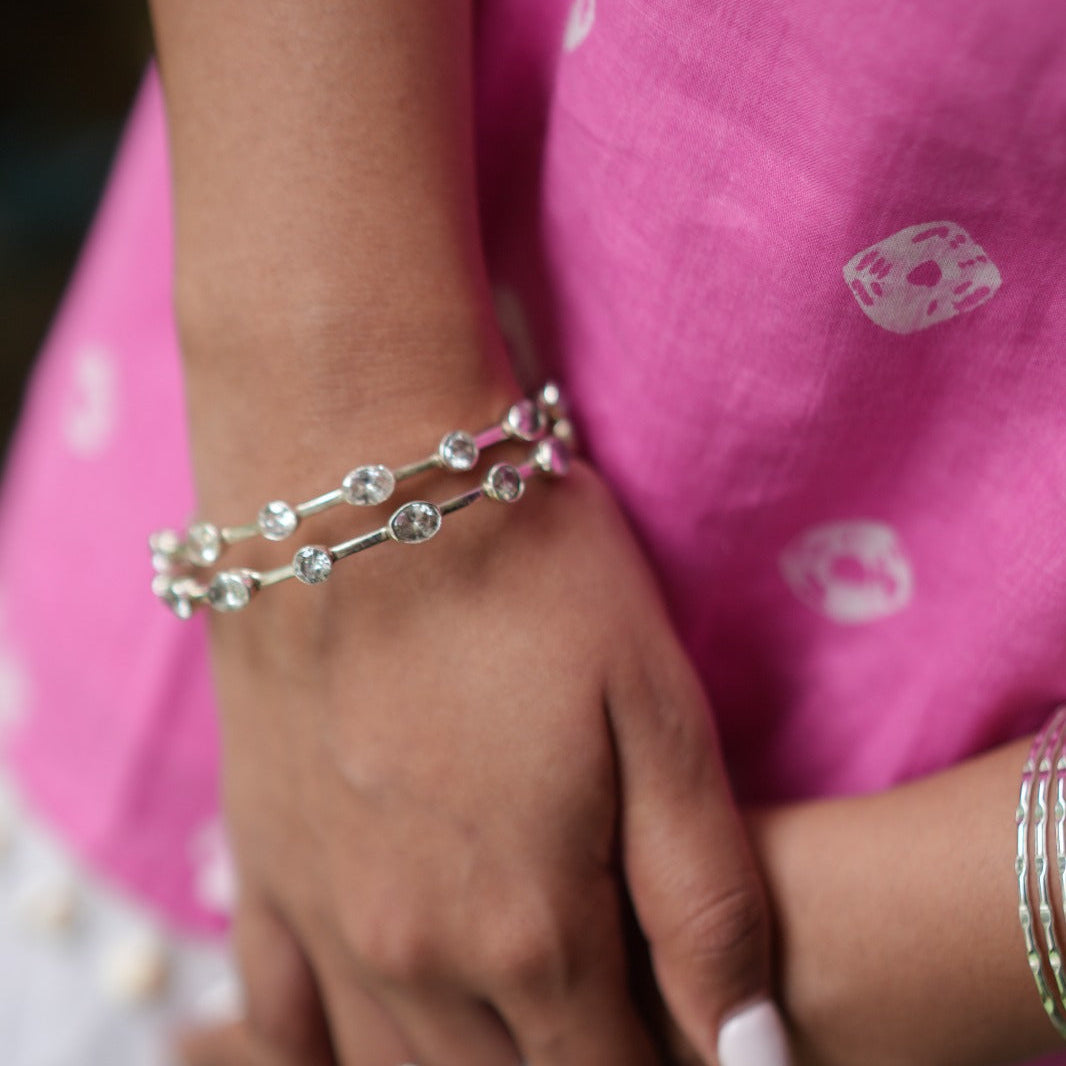 A close-up of a person's hands wearing a silver double-strand bracelet adorned with clear round crystals, along with a silver ring, set against a bright pink patterned garment.