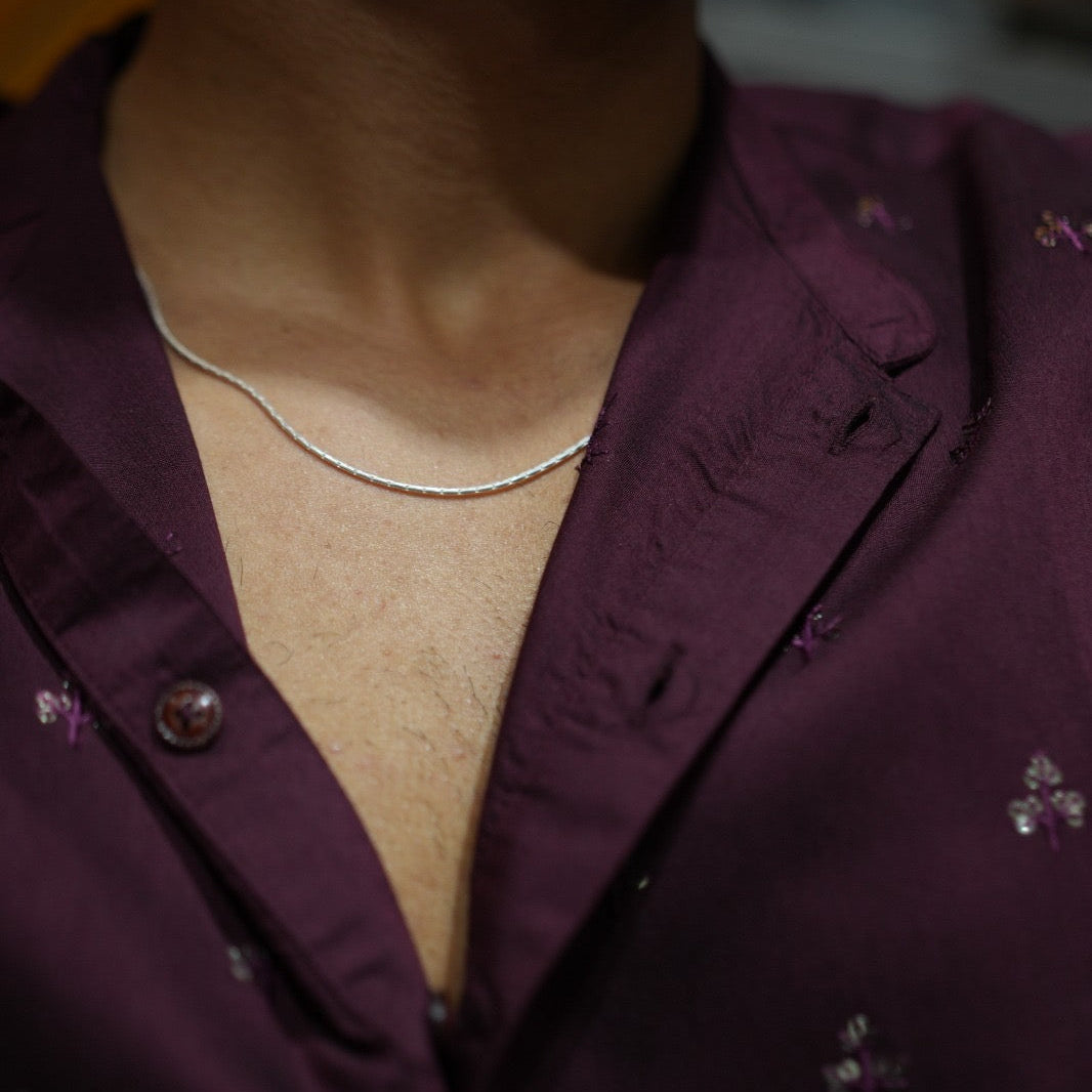 A close-up shot of a person wearing a thin silver chain necklace and a deep purple embroidered button-up shirt.